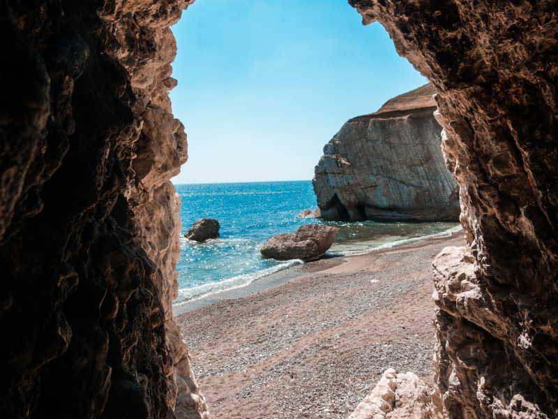 A view from inside a cave looking out to a pebble beach, blue sea, blue sky and rock formations