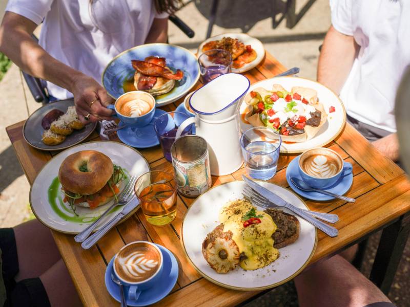 A wooden table outdoors is filled with plates of food and coffee
