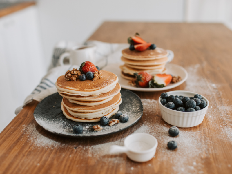 A dining table with two plates stacked high with pancakes. They are topped with berries and icing sugar