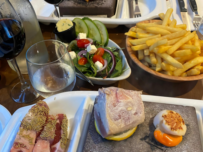 Meat on a hot stone and separate bowls of fries and Greek salad