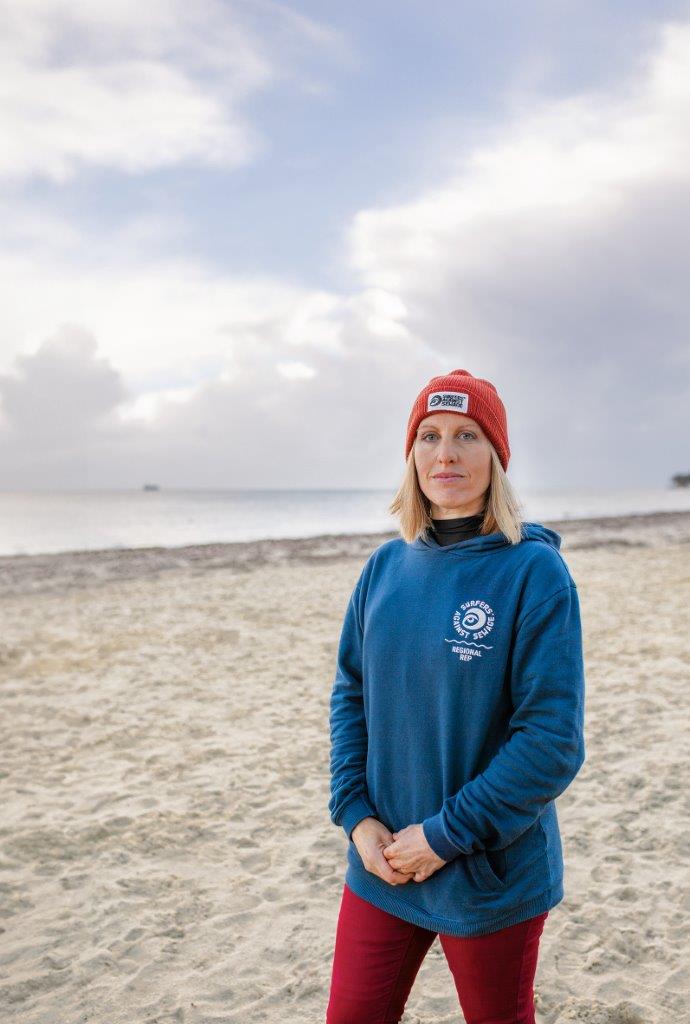 A woman with blonde shoulder-length hair standing on a sandy beach on the Isle of Wight, wearing a red beany hat and a blue sweatshirt