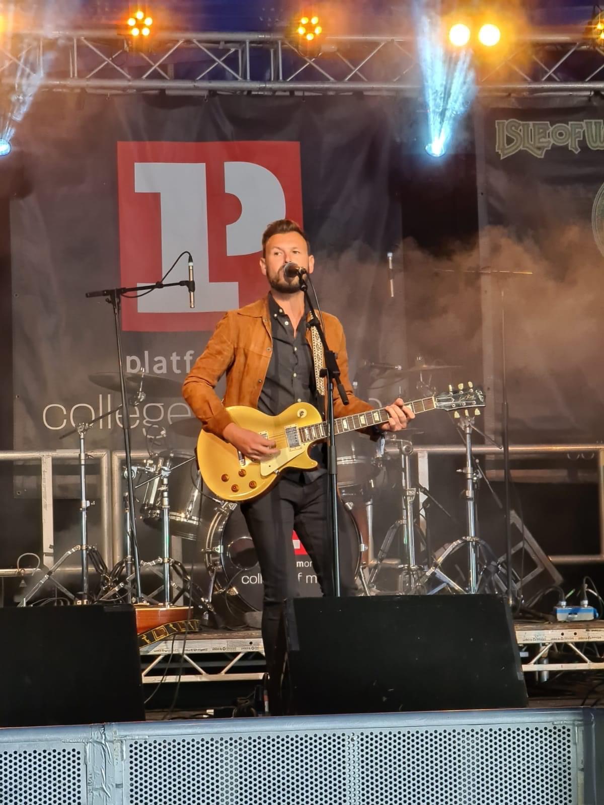 A man playing guitar and singing on a stage at a festival, he is wearing a tan jacket and dark clothing underneath