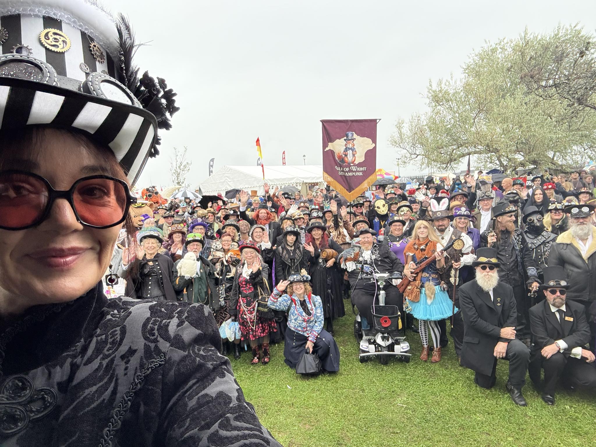Large group of people dressed in steampunk costumes posing for a photo at the Isle of Wight Steampunk Festival, with a participant in Victorian-inspired attire taking a selfie in the foreground.