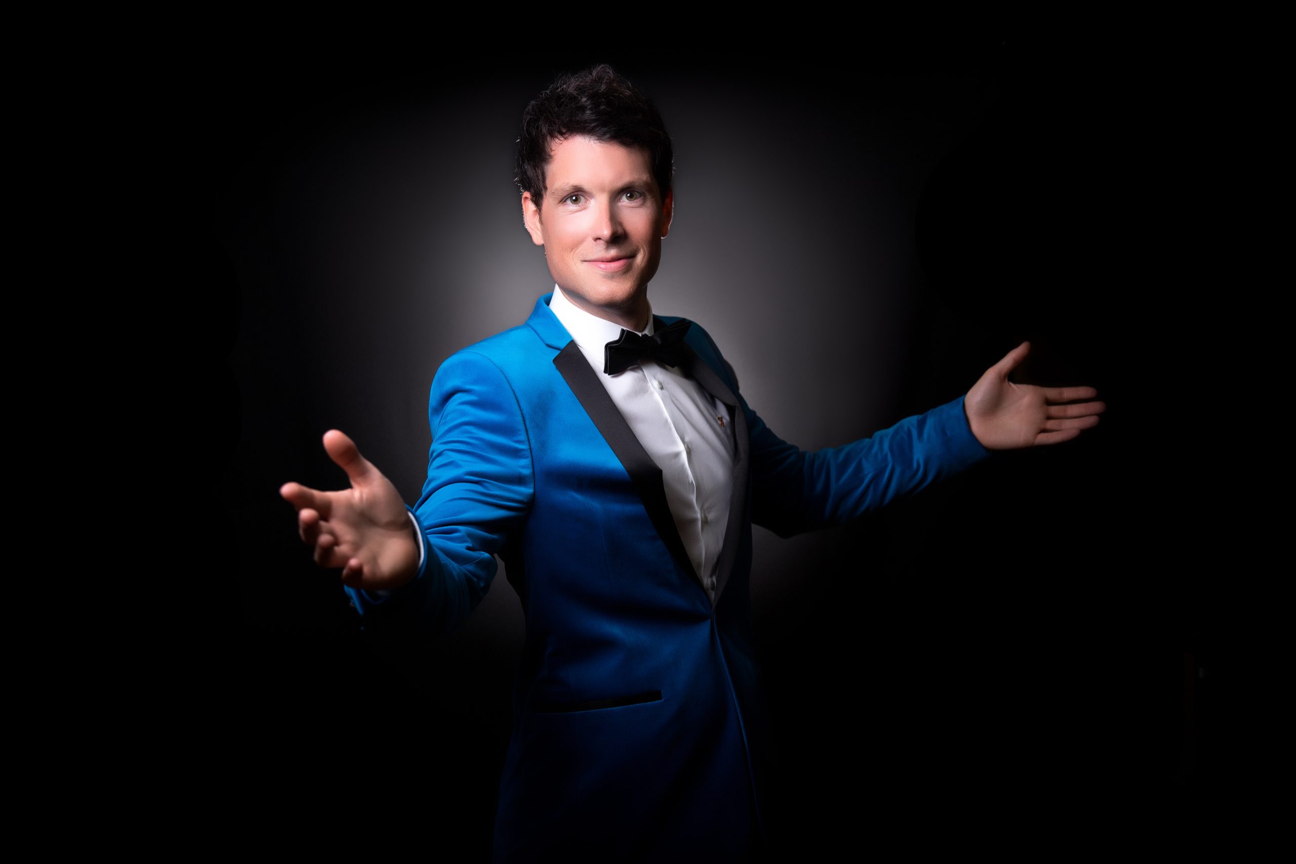 A singer - Mike Christi - in a spotlight wearing a blue tuxedo and black bow tie. His arms are outstretched.