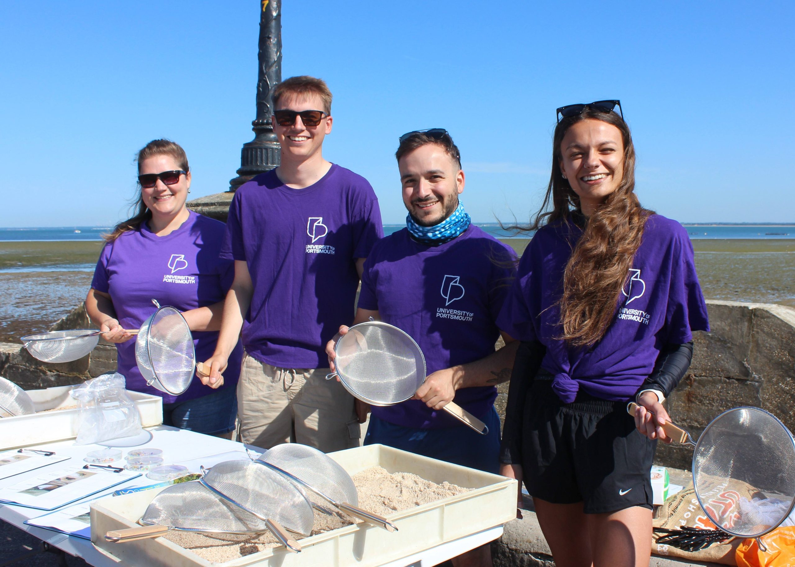 Four university of Portsmouth students in purple t-shirts stand by the beach with sieves for a scientific project