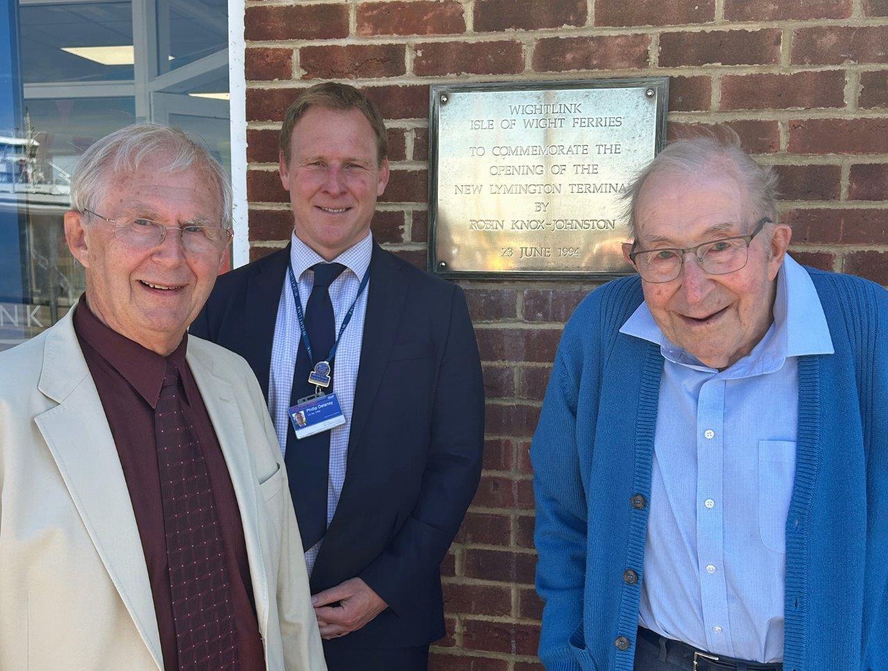 Three white men stand next to a gold-coloured plaque on an exterior brick wall