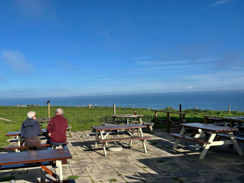 A patio with picnic tables on it, with two men sitting to the left, looks over green grass and down to the sea. It belongs to a pub.