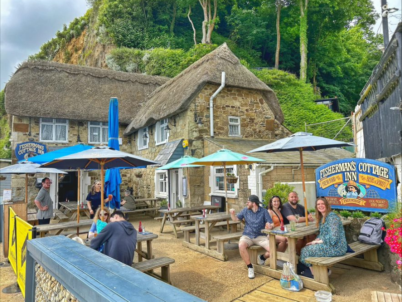 The exterior of a thatched English pub taken from the pub garden, with trees in the background. A couple of groups of friends sit under umbrellas at the wooden tables.