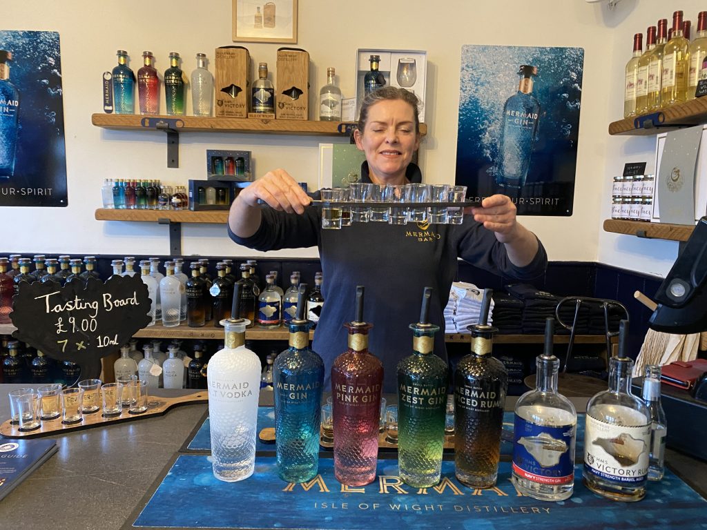 A woman holding up a tasting paddle of spirits made on the Isle of Wight. The corresponding bottles are on a bar in front of her.