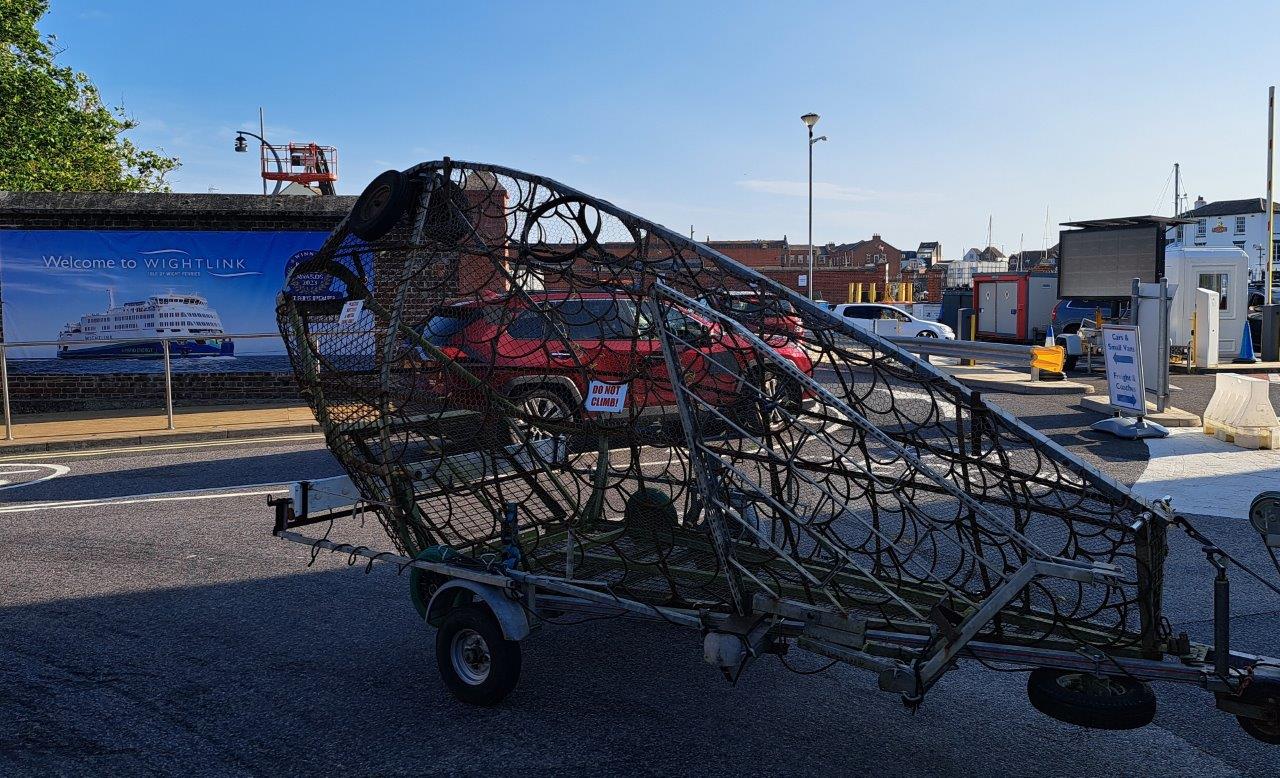 A sculpture made of metal wire in the shape of a fish is towed by a car on a trailer