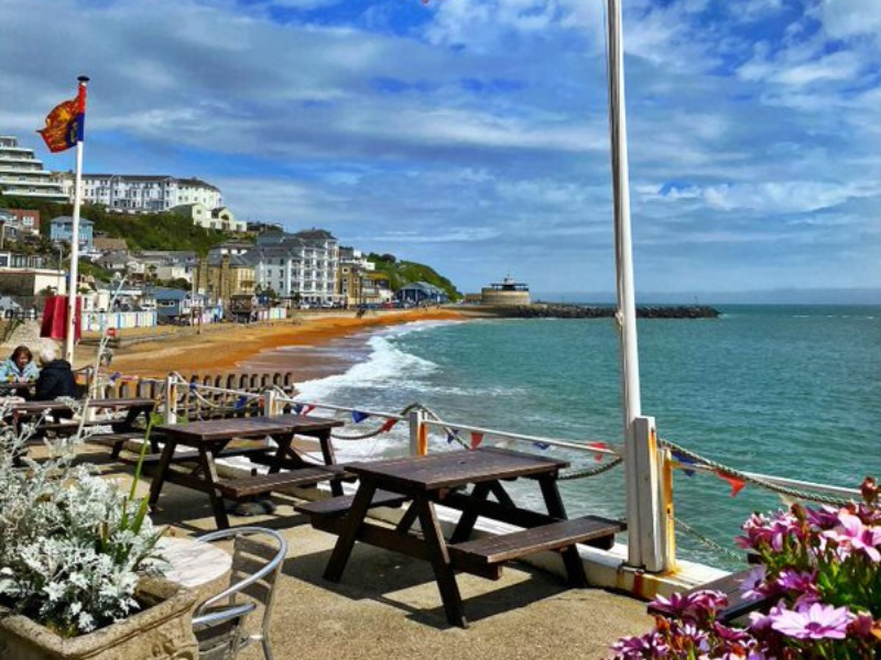 A photo of a beach taken from a pub's outside terrace. You can see wooden tables and plants in the foreground.