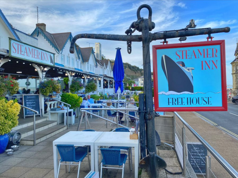 The exterior of a pub with the pub sign in shot, saying 'The Steamer Inn' with a picture of a steamer ship. You can see the pub's terrace with tables and chairs