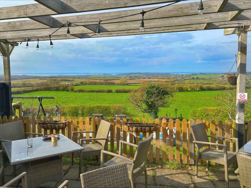 A country pub garden with views over English countryside and fields. Wooden chairs and tables sit beneath wooden beams adorned with festoon lighting.