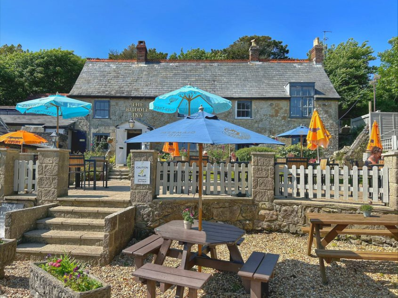 An old historic pub exterior, the photo taken from the pub garden with steps, a white fence and tables with umbrellas in the foreground