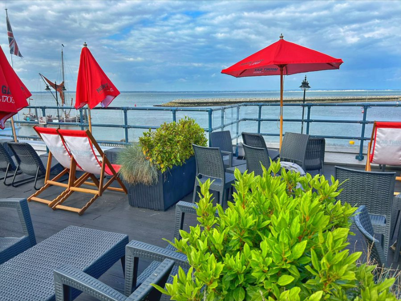 A terrace of a pub overlooking the sea. The terrace is dark wood and has deck chairs, red umbrellas and plants in pots