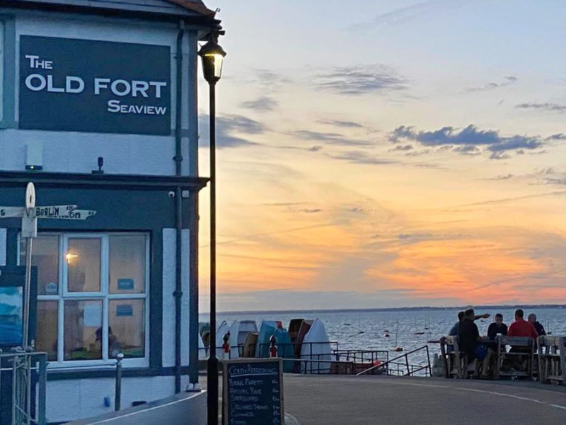 A white and blue pub with the sign 'The Old Fort' is to the left of a sea view with a sunset and people sat on a terrace