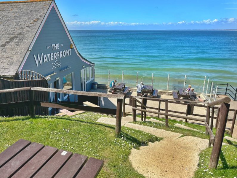 A pub called The Waterfront is pictured against a backdrop of blue sea. The photo is taken from a beer garden which has several levels and wooden tables.