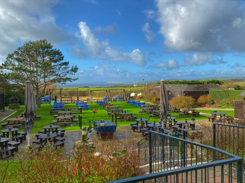 An extensive pub garden with terrace and grass is shown in the summer with blue sky. There are many tables for dining outdoors.