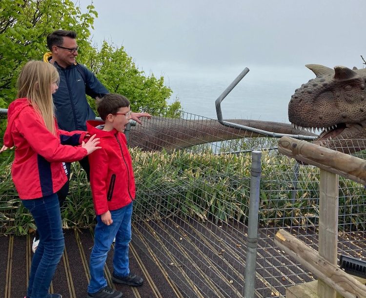 A family - man, woman and child - stand looking at a dinosaur exhibit at an outdoor attraction. It is overlooking the sea, the day is overcast and looks like its been raining