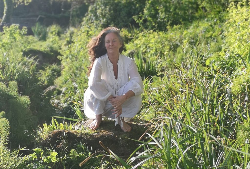 A woman with long brown hair and dressed in white linen is crouched on a rock amid some grass