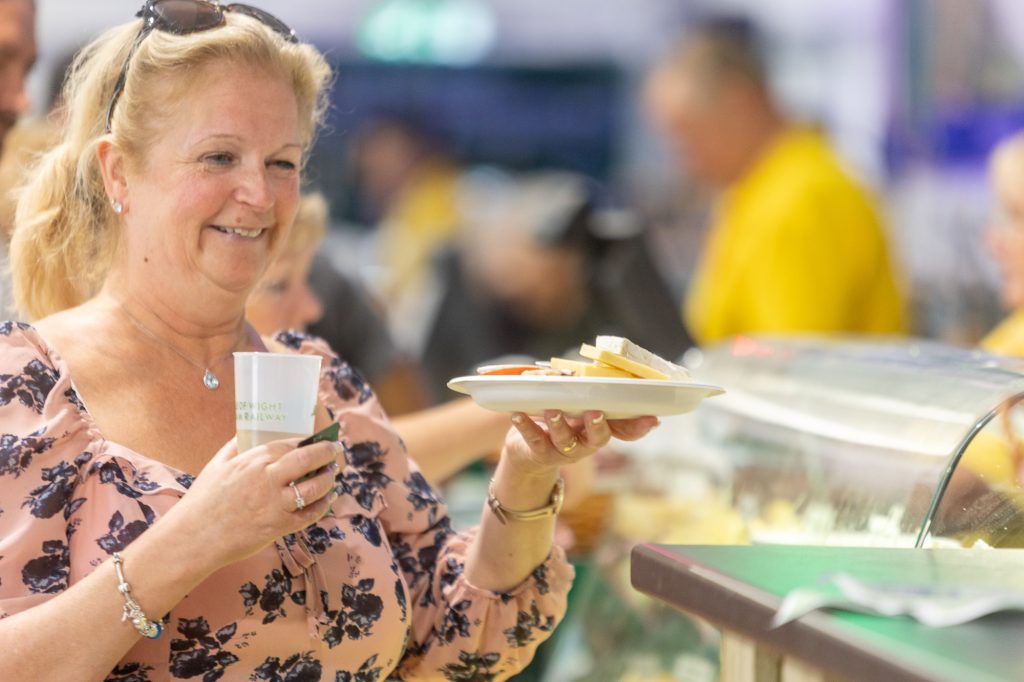 A smiling blonde woman holds a plate of cheese and a glass of cider at a stall at a festival