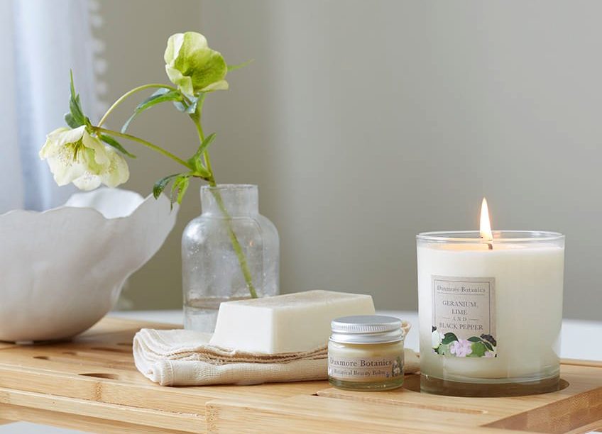 A lit candle, some body balm in a small jar and a bar of soap are on a wood worktop, with a flower in a vase