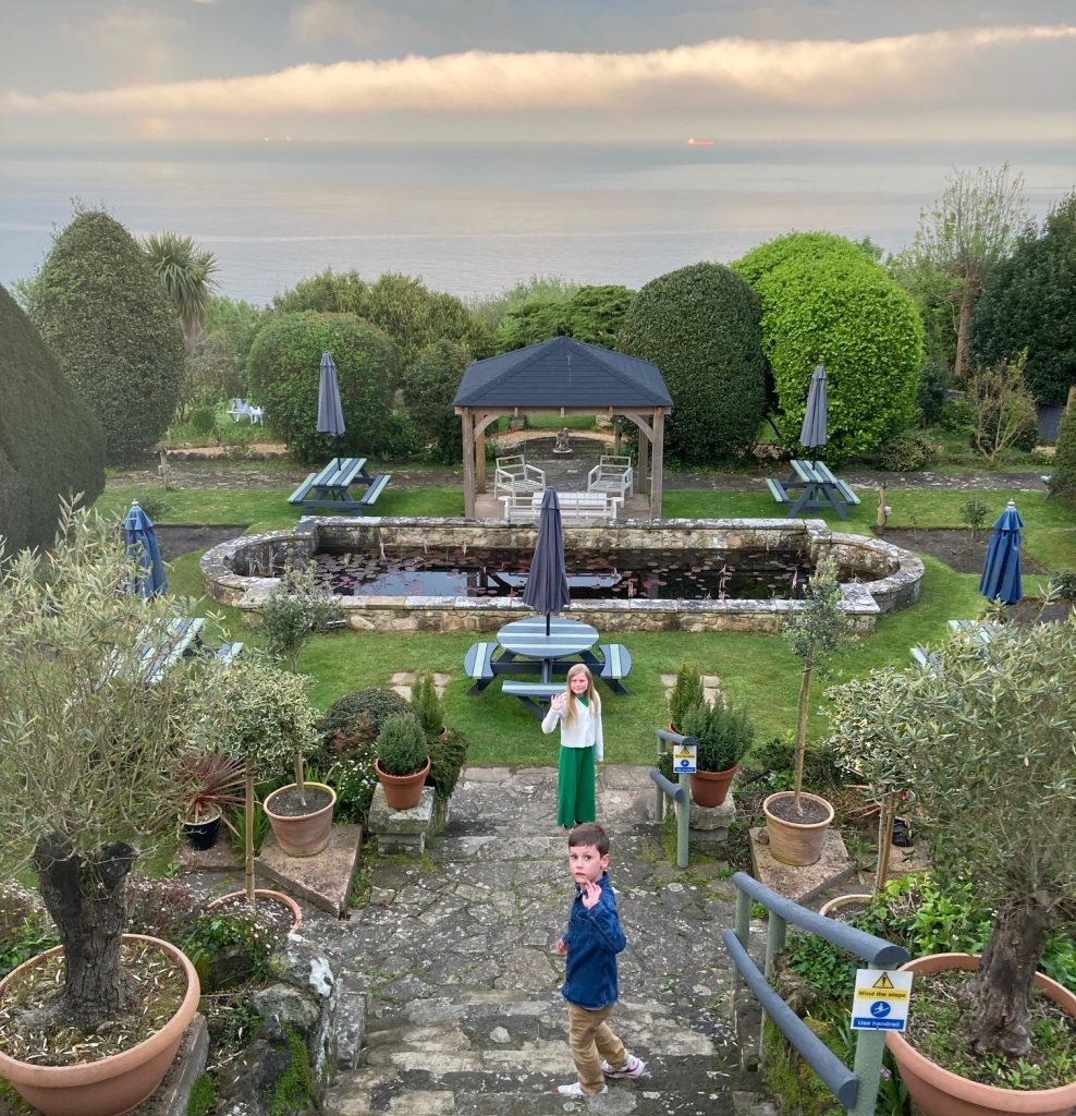 A view of a hotel garden with the photographer standing above it on some steps. On the stairs below is a boy and a woman waving. The garden has a pond