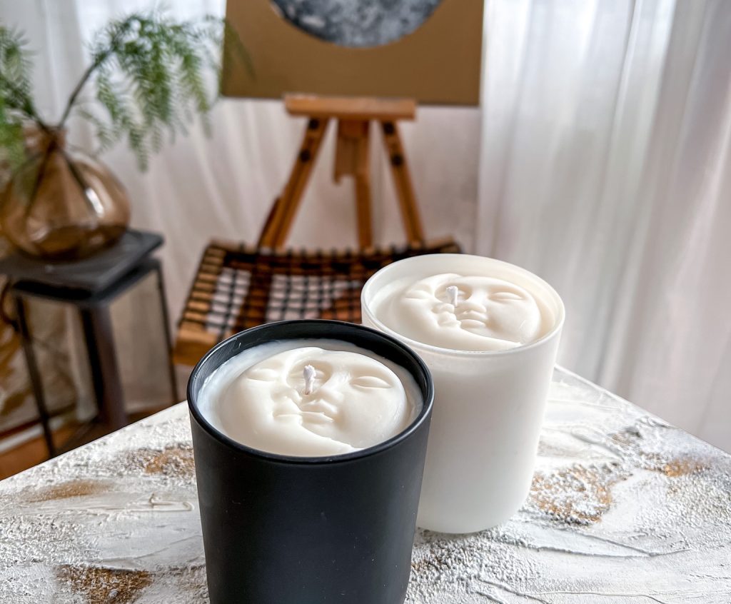 Two candles in glass jars, one black and one white, with faces embossed on them. There's a painting and a window in the background.