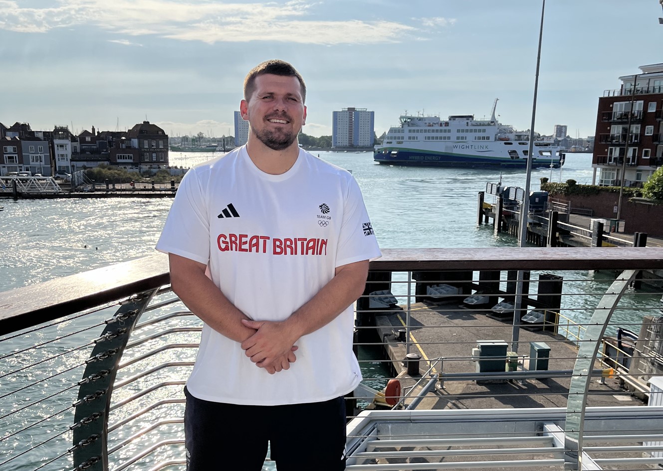 A man wearing a white Team GB t-shirt stands by a harbour with a Wightlink ferry in the background