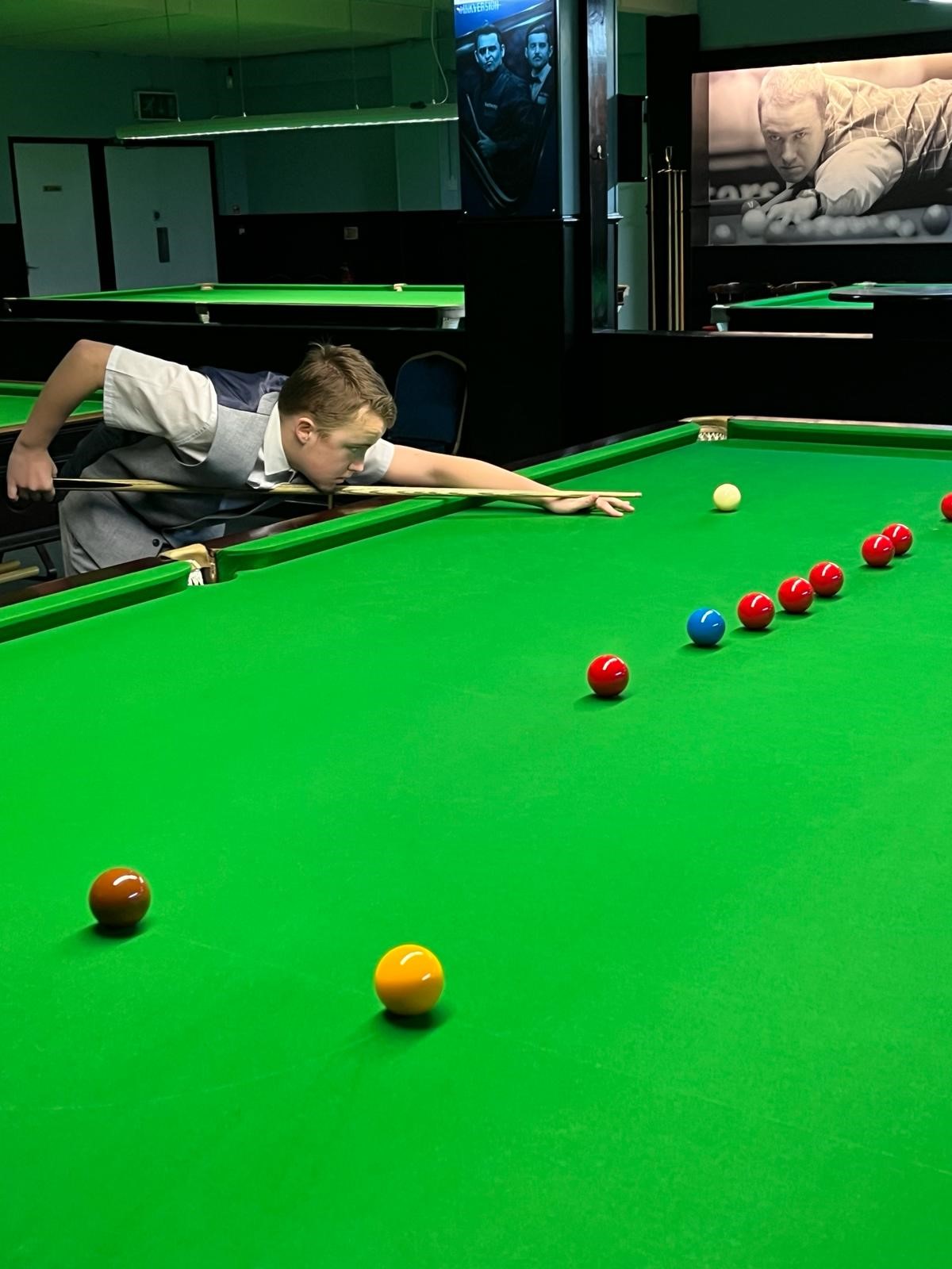 A snooker table with the classic green baize and various coloured balls on the table. A young player is leaning over the table taking a shot.