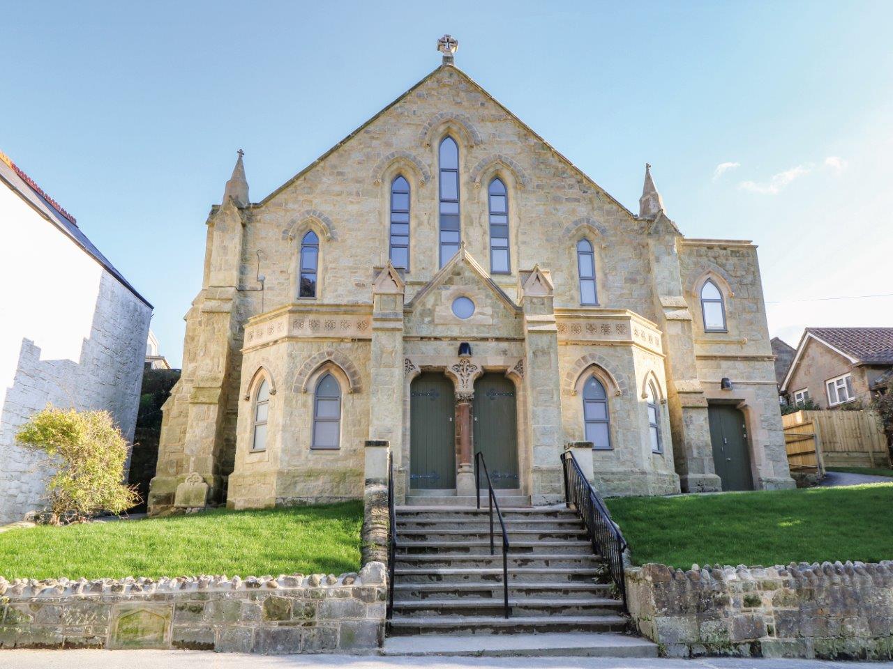 The exterior of a former church made from light coloured materials. There are steps to the font.