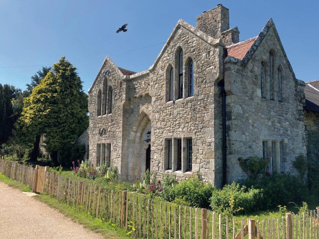 A sturdy stone cottage with a red roof, it's an Old Abbey Farmhouse. There's a fenced garden in front of it and a bird flying in the blue sky.