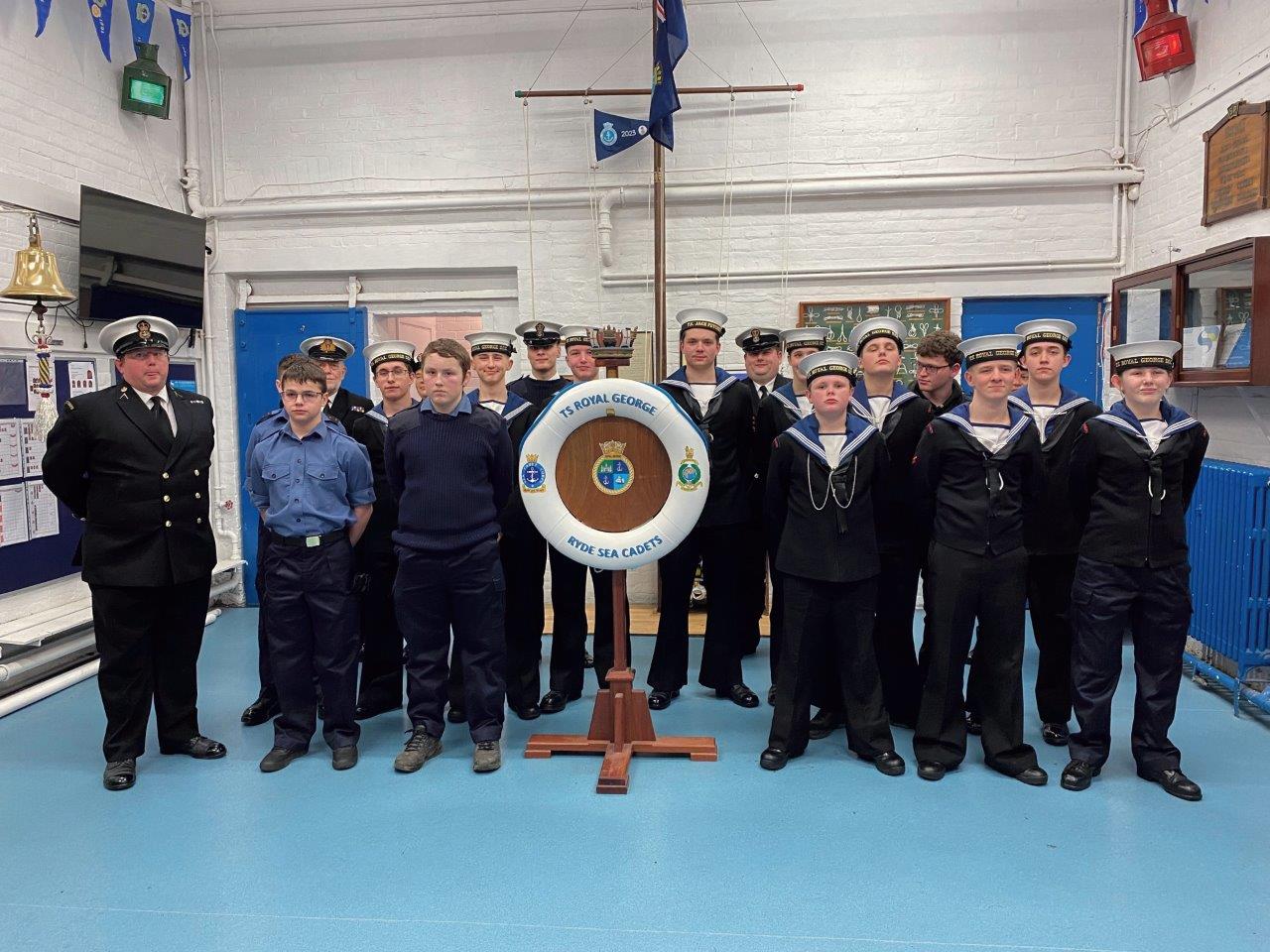 A group of youngsters stand inside a hall with a branded life ring, they are in Sea Cadet uniforms.