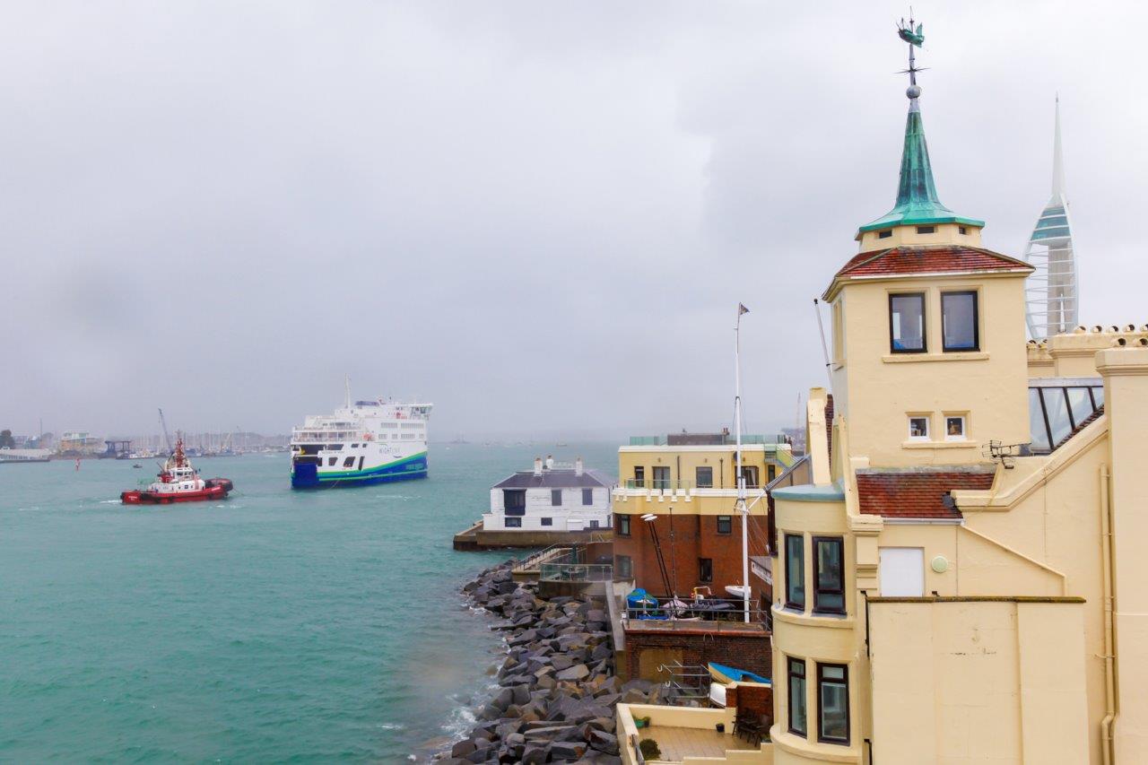 A scene of a historic city by the sea with boats on the water. We can see a turreted building on the shore and a ferry on the water