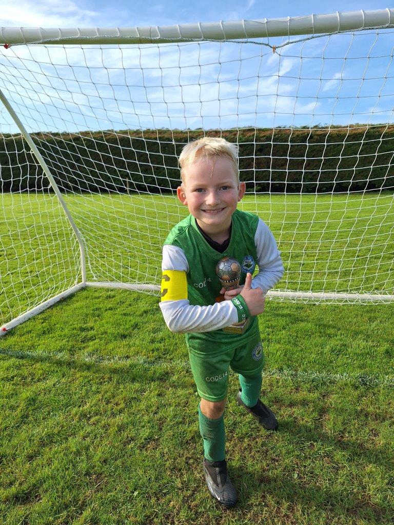 A child wearing a green football goalkeeper's outfit is posing for the camera with a trophy and a big smile inside an outdoor football net