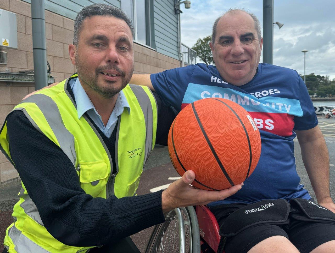 Two men, one using a wheelchair and one wearing a high visibility jacket, pose with a basketball.