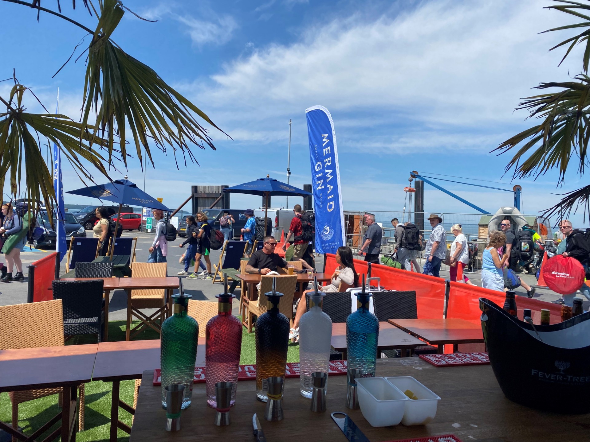 A view from behind a bar outdoors, with bottles of spirits and palm trees in the foreground, people and tables further away