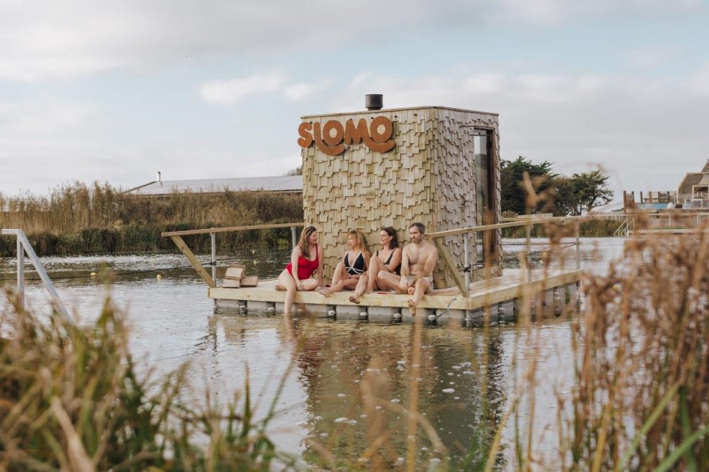 A sauna floating on a wooden platform on a lake, with countryside behind. There are people sat outside it overlooking the lake