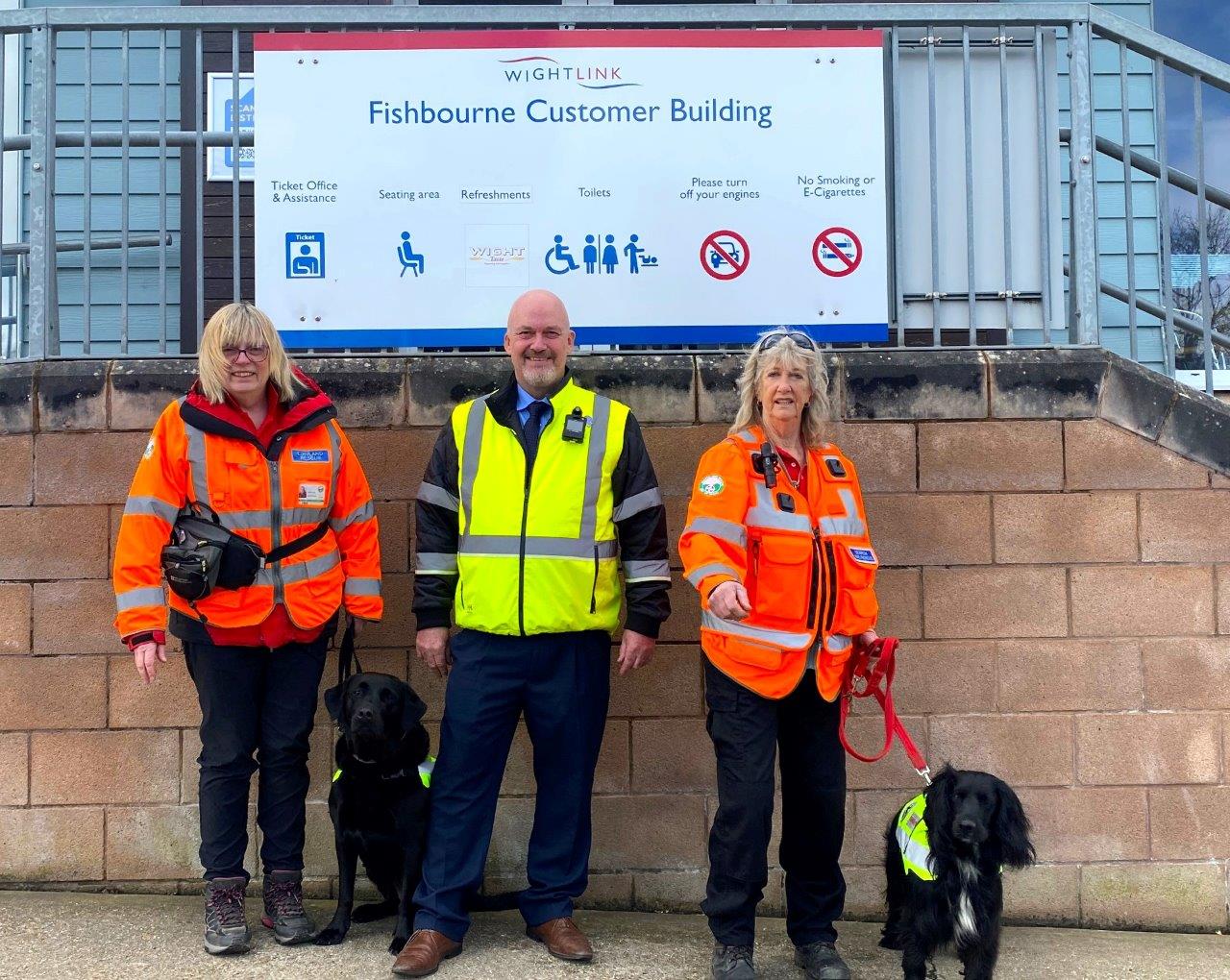 Two women wearing orange high visibility jackets stand with a man wearing a yellow high visibility jacket and two rescue dogs in front of a Wightlink ferries sign