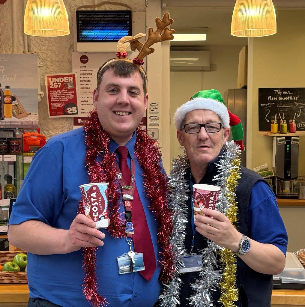 Two men, wearing blue Wightlink ferry uniforms, are draped in tinsel and wearing festive hats, holding coffee