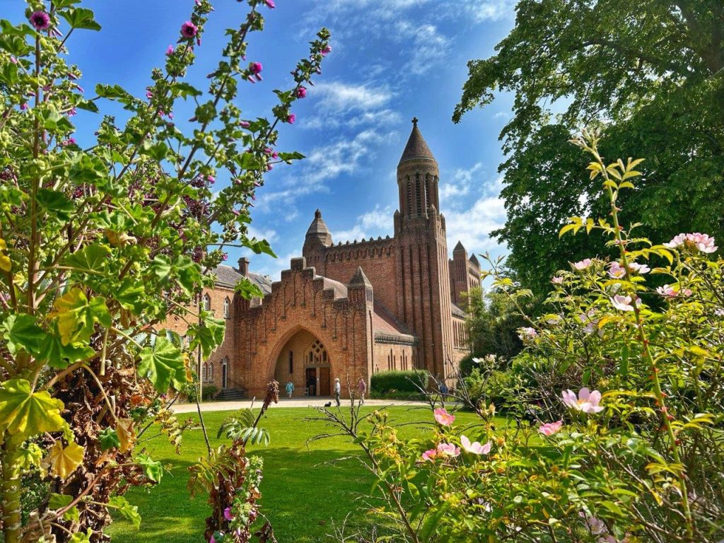 A monastery made of red brick and with intricate design is framed by grass, trees and garden