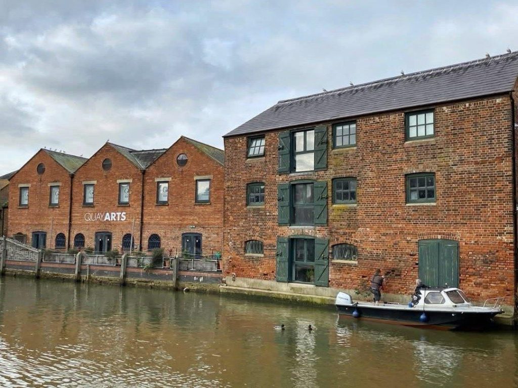 A red brick building housing an arts centre sits on a riverbank in a town setting