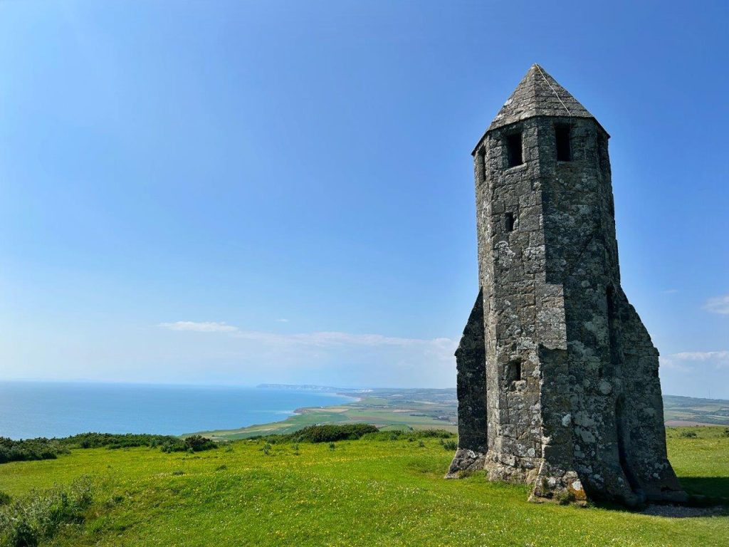 A tall, octagonal tower, the shell of a medieval lighthouse, sits on a green hill overlooking the sea