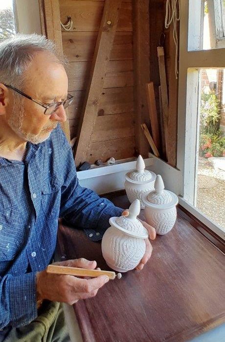 A man is at a wooden studio desk and has some pottery in his hands