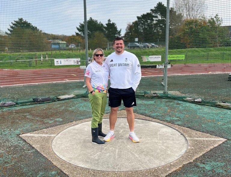 A man and a woman - mother and son - are standing together in a discuss circle in a sports field