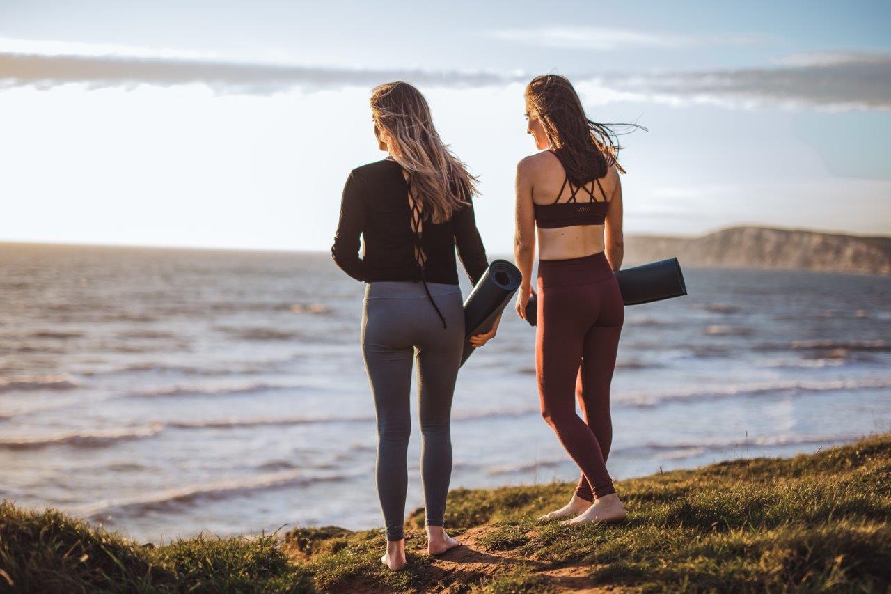 Two women walking towards the ocean. They are dressed in fitness outfits and carrying yoga mats