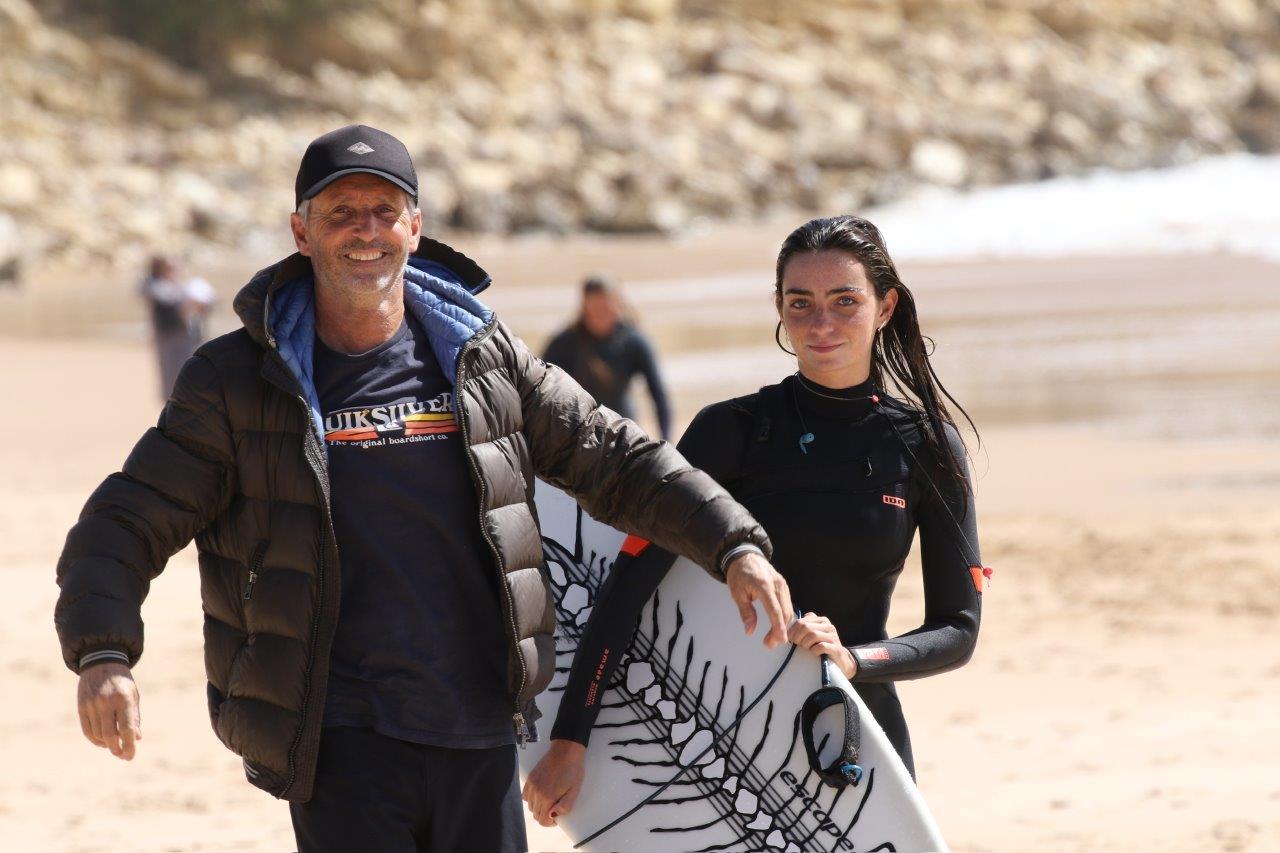 A man and a young woman (father and daughter) are on a sandy beach, standing together and smiling. The woman is holding a surf board and is in a wet suit