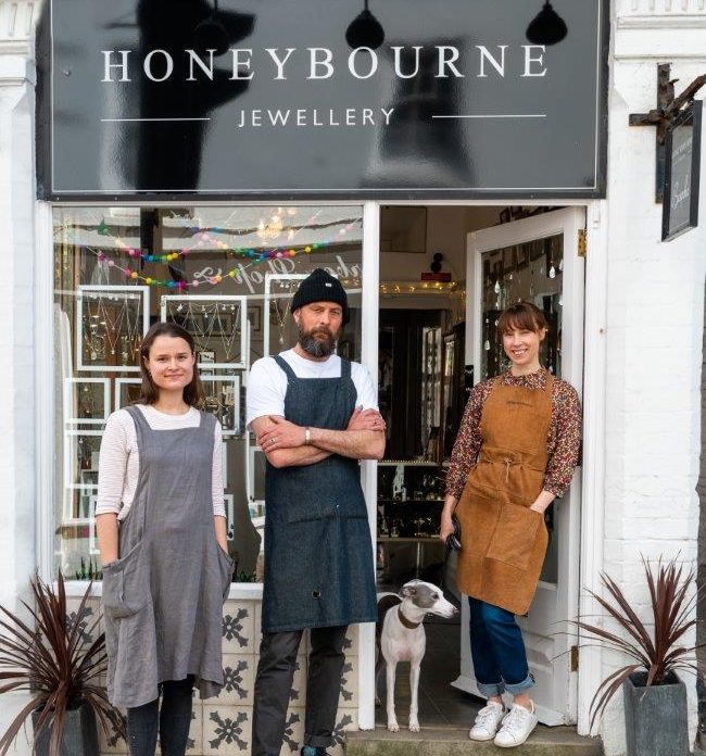 Three people wearing aprons and a dog stand outside a jewellers which has the sign Honeybourne Jewellery