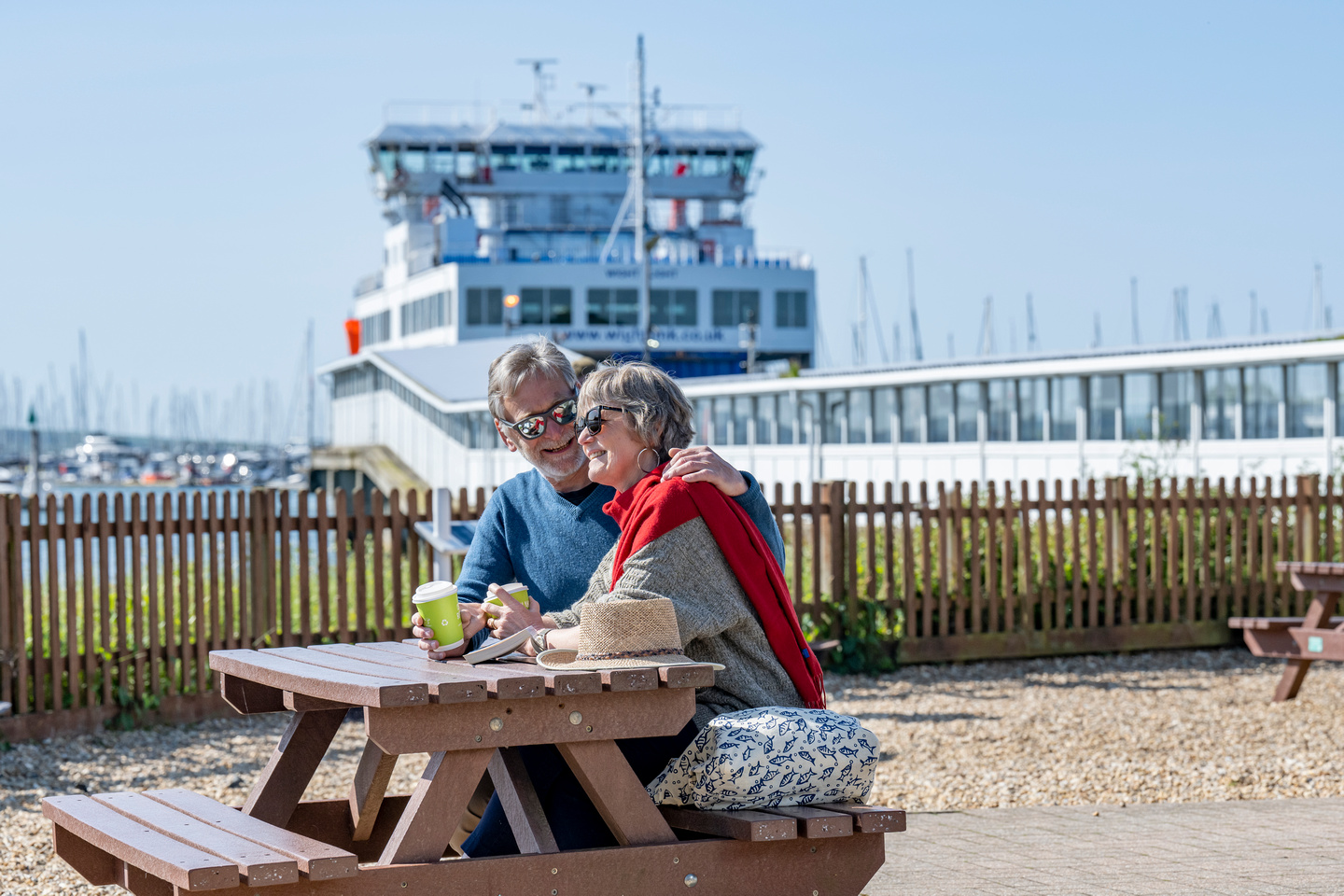 A couple sit at a bench at Wightlink's Lymington port, holding coffee cups and embracing. A ferry is in the background.