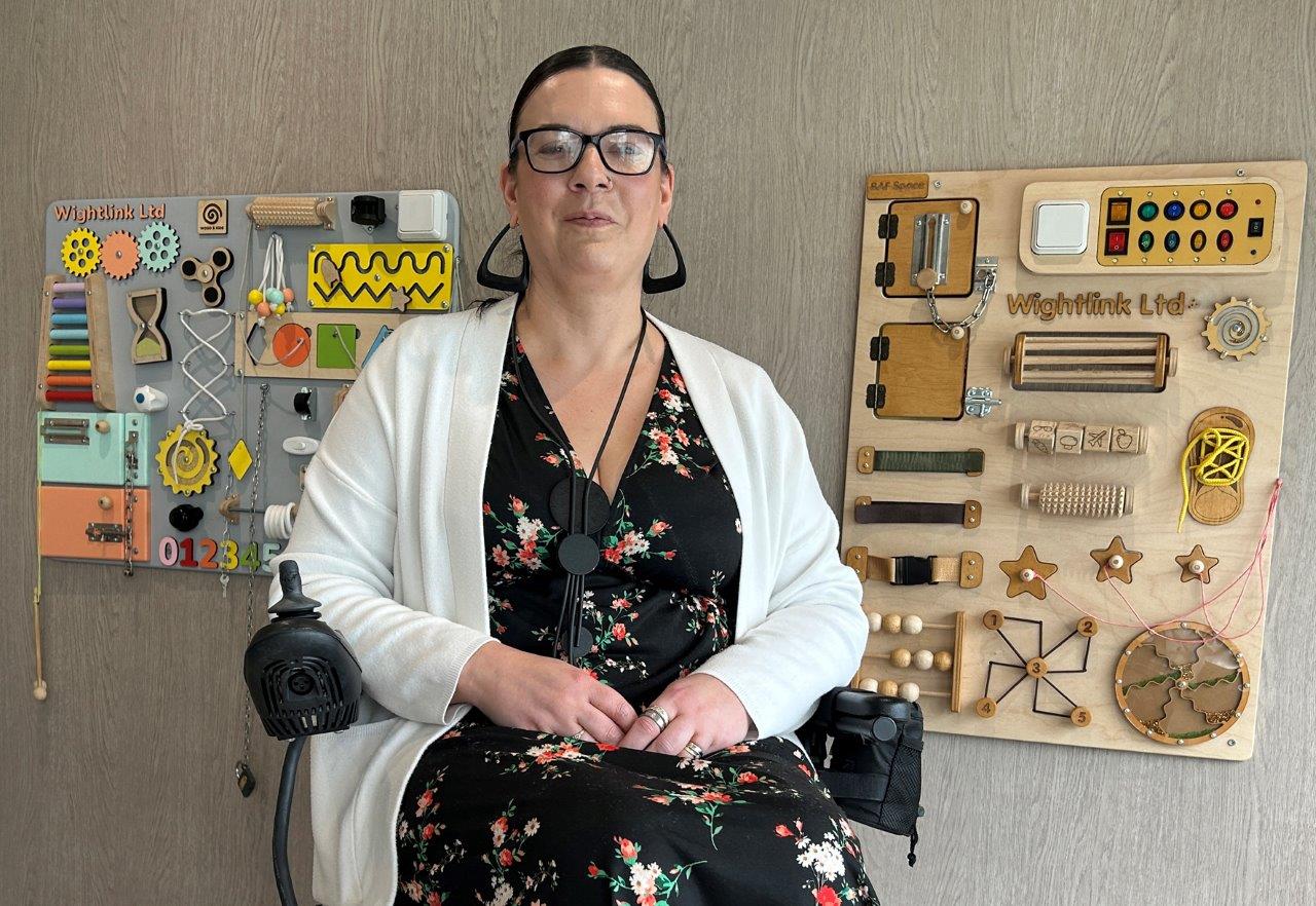 A white woman, using a wheelchair, sits in front of sensory boards.
